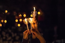 Several hands holding lit candles with protective covers in a dark setting. The warm glow of the candlelight illuminates the scene, creating a peaceful and solemn atmosphere. Blurred points of light in the background suggest other candles or light sources.