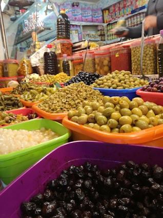 An array of colorful plastic bowls and containers filled with a variety of pickled and preserved foods, including green and black olives, onions, and other pickled vegetables. The background shows a cluttered market stall with various jars and packages stacked on shelves. A person is visible in the background holding an item.