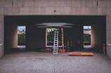 A construction or maintenance area with a tall ladder positioned under a circular structure inside a building with a tiled surface. Orange and yellow safety barriers lie on the ground nearby. The setting suggests an area undergoing renovation or installation work, with a partially hidden person and some construction materials visible in the background.