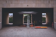 A construction or maintenance area with a tall ladder positioned under a circular structure inside a building with a tiled surface. Orange and yellow safety barriers lie on the ground nearby. The setting suggests an area undergoing renovation or installation work, with a partially hidden person and some construction materials visible in the background.
