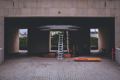 A construction or maintenance area with a tall ladder positioned under a circular structure inside a building with a tiled surface. Orange and yellow safety barriers lie on the ground nearby. The setting suggests an area undergoing renovation or installation work, with a partially hidden person and some construction materials visible in the background.