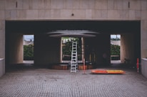 A construction or maintenance area with a tall ladder positioned under a circular structure inside a building with a tiled surface. Orange and yellow safety barriers lie on the ground nearby. The setting suggests an area undergoing renovation or installation work, with a partially hidden person and some construction materials visible in the background.