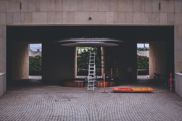 A construction or maintenance area with a tall ladder positioned under a circular structure inside a building with a tiled surface. Orange and yellow safety barriers lie on the ground nearby. The setting suggests an area undergoing renovation or installation work, with a partially hidden person and some construction materials visible in the background.
