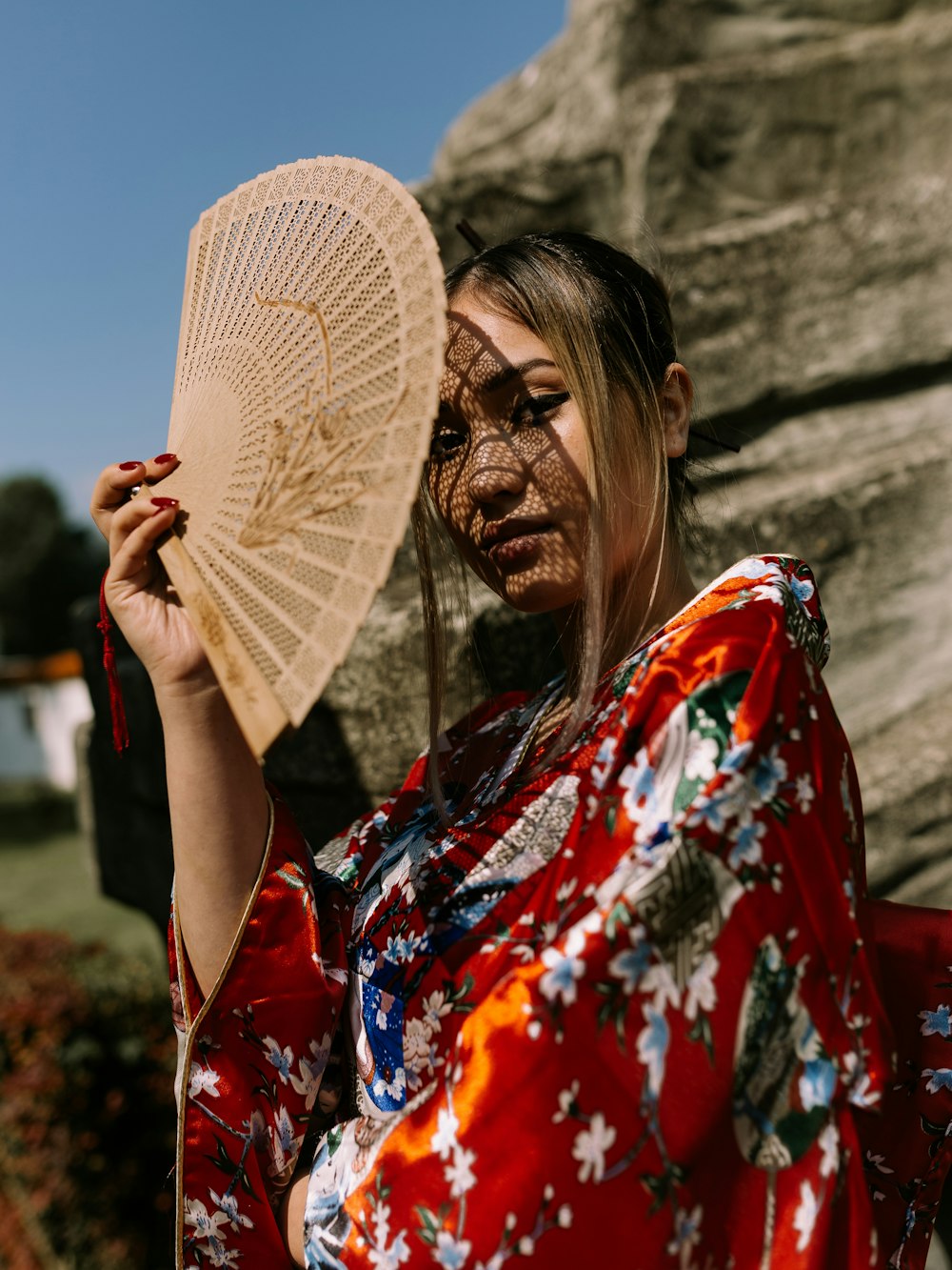 Femme en kimono floral rouge, blanc et bleu debout et couvrant son visage avec un éventail en bois