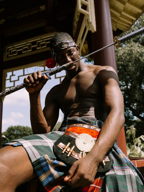 A shirtless person wearing a kilt is sitting with a katana held to their mouth. There is a red rose on the katana. The background includes a structure with traditional Asian architectural elements and trees.