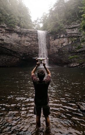 A person stands in shallow water, holding a drone aloft, in front of a large, cascading waterfall surrounded by forested cliffs.
