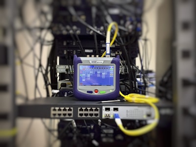 A network tester with a screen displaying data is placed on a server rack, surrounded by cables and networking equipment.