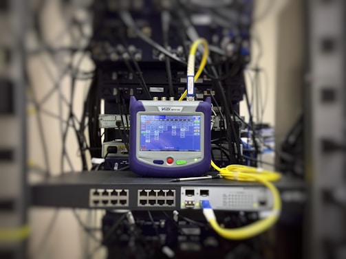 A network tester with a screen displaying data is placed on a server rack, surrounded by cables and networking equipment.