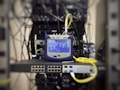 A network tester with a screen displaying data is placed on a server rack, surrounded by cables and networking equipment.