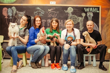 Five people are seated in front of a chalkboard with animal drawings and the word 'WAHL' visible. Each person is holding a different animal, including a fluffy white dog, a rooster, a hedgehog, and a small poodle. They are casually posing for the camera, with a cheerful demeanor. The chalkboard in the background features drawings of balloons, a cat, rabbits, and a poodle.