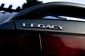 A close-up shot of a car's rear, featuring a shiny 'LEGACY' emblem. The background is slightly blurred, with a hint of trees or a structure visible.