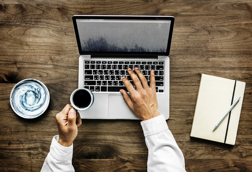 person using black and gray laptop computer while holding white ceramic mug with coffee in top view photography