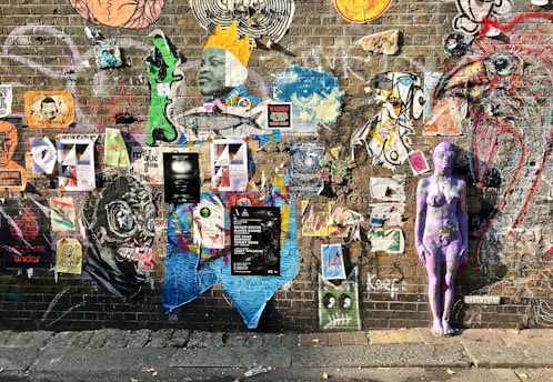 A vibrant brick wall adorned with a diverse array of street art, including posters, graffiti, and a painted purple human figure. The art varies in style and content, featuring faces, abstract designs, and large animal figures, conveying a sense of creativity and urban expression.