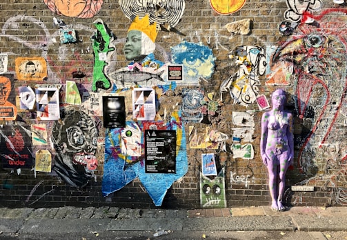A vibrant brick wall adorned with a diverse array of street art, including posters, graffiti, and a painted purple human figure. The art varies in style and content, featuring faces, abstract designs, and large animal figures, conveying a sense of creativity and urban expression.