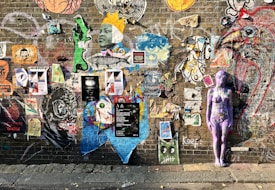 A vibrant brick wall adorned with a diverse array of street art, including posters, graffiti, and a painted purple human figure. The art varies in style and content, featuring faces, abstract designs, and large animal figures, conveying a sense of creativity and urban expression.