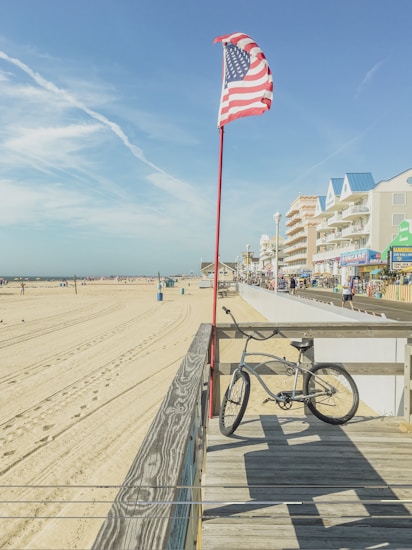 A bicycle is leaning against a wooden railing on a boardwalk near a sandy beach. An American flag is waving on a tall red pole nearby. The beach extends into the distance, with footprints and tire tracks visible in the sand. Behind the boardwalk, there are several buildings with bright signage, likely a coastal town.