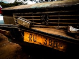 A close-up of the front of an old, weathered car with a prominent grille. The vehicle has a distinctive emblem on the grille and a yellow license plate with the characters 'RU 380'. The car's surface is rusty and dirty, giving an impression of age and disuse.