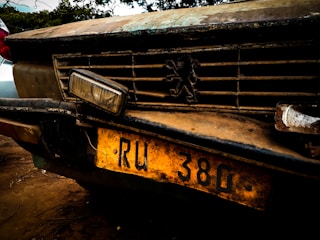 A close-up of the front of an old, weathered car with a prominent grille. The vehicle has a distinctive emblem on the grille and a yellow license plate with the characters 'RU 380'. The car's surface is rusty and dirty, giving an impression of age and disuse.