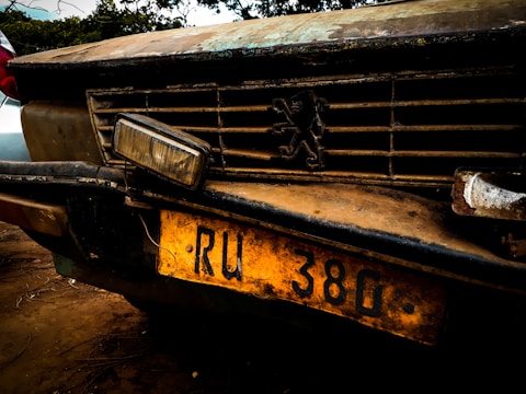 A close-up of the front of an old, weathered car with a prominent grille. The vehicle has a distinctive emblem on the grille and a yellow license plate with the characters 'RU 380'. The car's surface is rusty and dirty, giving an impression of age and disuse.