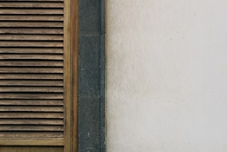 A portion of a building wall featuring a wooden louvered panel on the left. The panel has vertical slats with a natural wood finish. Next to the panel is a section of dark gray stone, followed by a pale, slightly weathered and cracked plaster wall on the right.