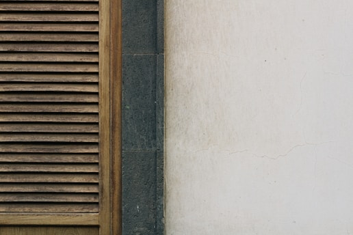 A portion of a building wall featuring a wooden louvered panel on the left. The panel has vertical slats with a natural wood finish. Next to the panel is a section of dark gray stone, followed by a pale, slightly weathered and cracked plaster wall on the right.