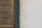 A portion of a building wall featuring a wooden louvered panel on the left. The panel has vertical slats with a natural wood finish. Next to the panel is a section of dark gray stone, followed by a pale, slightly weathered and cracked plaster wall on the right.