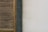 A portion of a building wall featuring a wooden louvered panel on the left. The panel has vertical slats with a natural wood finish. Next to the panel is a section of dark gray stone, followed by a pale, slightly weathered and cracked plaster wall on the right.