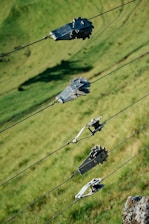 Several metal gears and clamps are attached to tensioned wires positioned diagonally across the image. The background is a grassy hillside with varying shades of green, providing a natural setting.