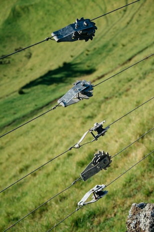 Several metal gears and clamps are attached to tensioned wires positioned diagonally across the image. The background is a grassy hillside with varying shades of green, providing a natural setting.