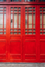 A close-up of a vibrant red wooden door with decorative grid patterns and frosted glass panels. The door features a mix of vertical and horizontal wooden slats, creating a geometric design. The lower section of the door is solid wood, while the upper section is divided by a series of small glass panes.