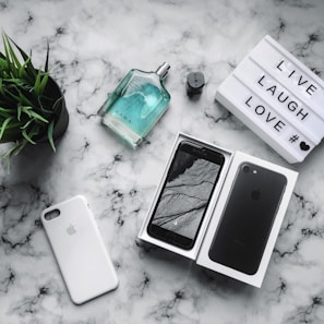 A flat lay arrangement on a marble surface includes a smartphone in a box, an empty phone case, a bottle of Zara perfume, a potted plant, and a lightbox with the words 'LIVE LAUGH LOVE'. The items are neatly organized, creating a stylish and modern aesthetic.