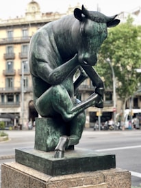 A bronze statue of a bull seated in a contemplative pose, reminiscent of Rodin's 'The Thinker.' The sculpture is set on a stone pedestal in an urban setting with a blurred background of buildings and trees.