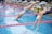 Several swimmers in competition dive into a swimming pool. They are wearing colorful swimwear and caps, and are in the midst of an athletic event, with droplets of water splashing as they enter the water. The scene captures the motion and excitement of the race.