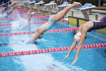 Several swimmers in competition dive into a swimming pool. They are wearing colorful swimwear and caps, and are in the midst of an athletic event, with droplets of water splashing as they enter the water. The scene captures the motion and excitement of the race.