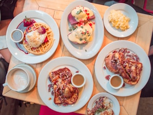 A wooden table is filled with a variety of breakfast dishes, including waffles with whipped cream and strawberries, eggs Benedict topped with hollandaise sauce and garnished with herbs, scrambled eggs, and plates of French toast with pecans and syrup. A cup of cappuccino with latte art is also visible.