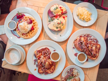 A wooden table is filled with a variety of breakfast dishes, including waffles with whipped cream and strawberries, eggs Benedict topped with hollandaise sauce and garnished with herbs, scrambled eggs, and plates of French toast with pecans and syrup. A cup of cappuccino with latte art is also visible.