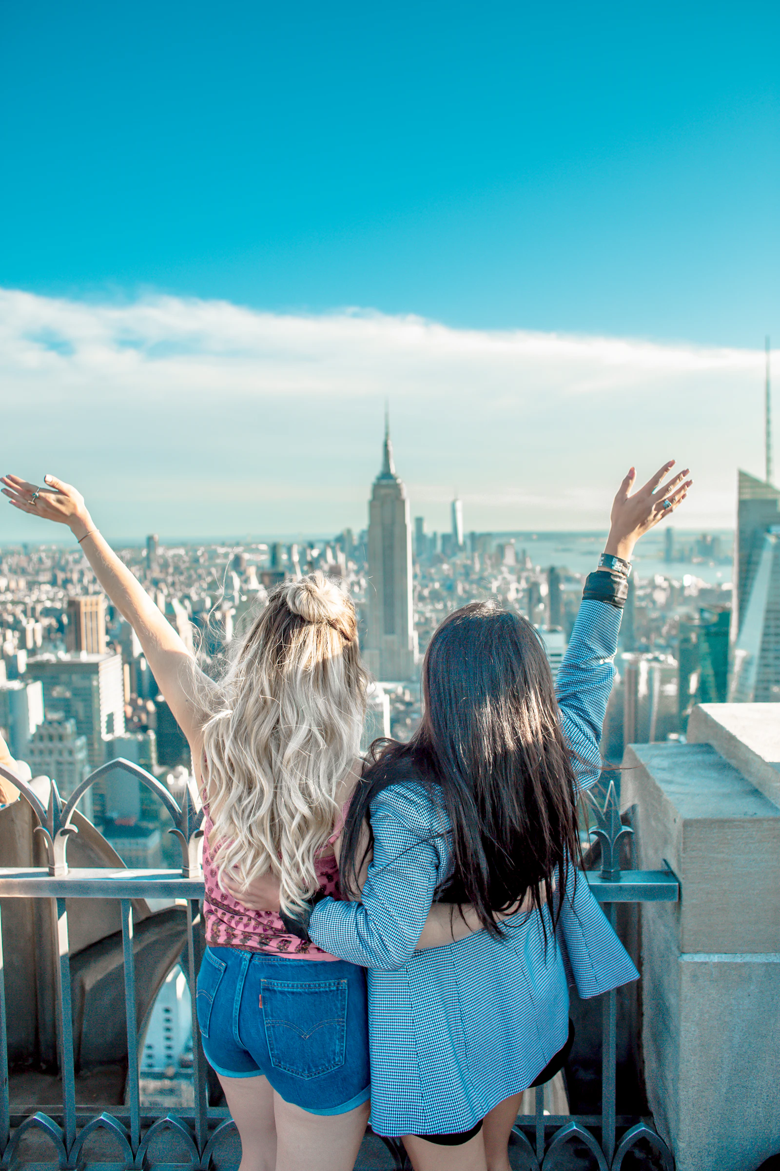 Two travellers stand together at a high city viewpoint with their arms raised towards the skyline.