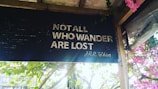 A sign with the quote 'Not all who wander are lost' by J.R.R. Tolkien is displayed. The sign is dark with white lettering and is surrounded by a lush setting with green leaves and pink flowers.