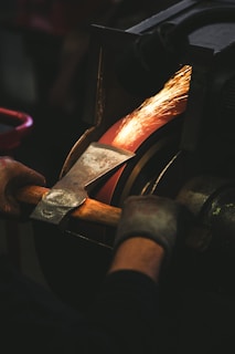 A person wearing gloves is sharpening an axe blade on a grinding wheel, with bright sparks flying off as the metal is honed.