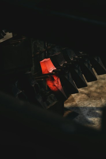 A glowing piece of metal is clamped between metal jaws, likely in a workshop or industrial setting. The metal appears to have been heated recently, as it emits a bright red-orange hue. The surrounding area is dimly lit, highlighting the contrast between the glowing metal and the dark machinery.