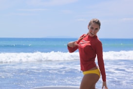 A woman standing on a beach in front of the ocean, wearing a red rash guard and yellow bikini bottom. She is smiling and pointing at something on her shirt. The sea is calm with gentle waves, and the sky is clear with a few clouds.