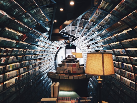 A library with a curved tunnel-like design featuring walls lined with bookshelves filled with books. The warm lighting and cozy atmosphere are enhanced by several large lamps placed on tables filled with more books.