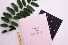 A pink notebook with the phrase 'Today I Am Grateful' is placed on a flat surface. Beside it, there is a black card with small heart patterns and mostly unreadable text. A gold pen lies next to the notebook. A branch with green leaves is also placed alongside, adding a natural element to the composition.