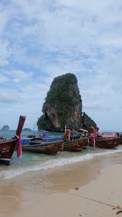 A sandy beach with several traditional longtail boats tied up along the shoreline, each adorned with colorful fabric. In the background, a large limestone rock formation covered with green foliage rises from the sea. The sky is mostly cloudy, creating a serene and relaxed atmosphere.