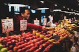 A fruit vendor is engaging with a customer at a busy market stall. Various types of fruit, including apples, pears, and mangoes, are displayed on the counter, with price tags visible. The vendor is handing over change to the customer, and there are overhead lights illuminating the vivid colors of the fruits.