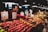 A fruit vendor is engaging with a customer at a busy market stall. Various types of fruit, including apples, pears, and mangoes, are displayed on the counter, with price tags visible. The vendor is handing over change to the customer, and there are overhead lights illuminating the vivid colors of the fruits.