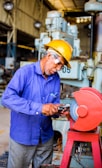 A man wearing a blue shirt, safety glasses, and a yellow hard hat operates a grinding machine. Sparks fly as he sharpens a tool in an industrial setting with large machinery in the background.