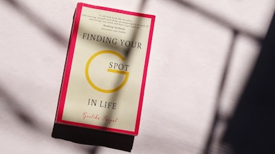 A book with a red border placed on a light-colored surface displaying the title 'Finding Your G Spot in Life' by Geetika Saigal. Sunlight creates shadows on the surface.