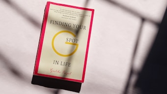 A book with a red border placed on a light-colored surface displaying the title 'Finding Your G Spot in Life' by Geetika Saigal. Sunlight creates shadows on the surface.