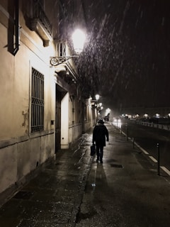 A cinematic shot of Saint Amari walking alone down a rain-soaked street at night, illuminated by distant city lights.